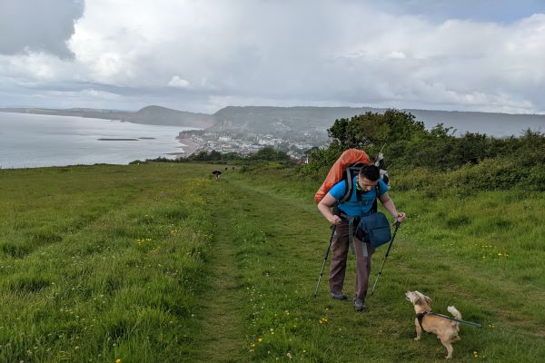 Ryan Eddowes is walking up a large grassy hill. The view of the red cliffs of Sidmouth extends into the background. Ryan is using waling poles to assist his walking and is carrying a large rucksack on his back. A small, sandy-coloured dog is looking up at him by his feet. The weather is misty and cloudy.